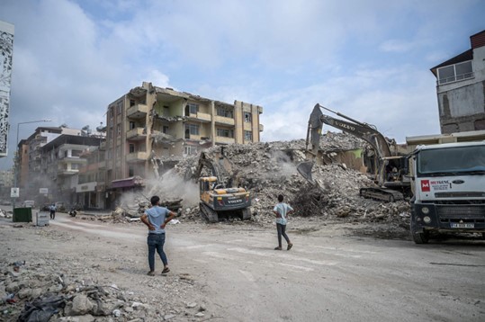 Orang-orang menyaksikan para penggali bekerja untuk membersihkan puing-puing bangunan yang runtuh, lima bulan setelah gempa bumi dahsyat melanda Turki tenggara, di Samandağ, Hatay, 9 Juli 2023. (Foto: AFP)